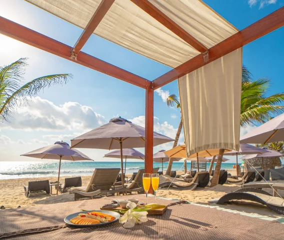 Shaded beach cabana at MVNGATA with drinks and a fruit plate, umbrellas and the sea behind