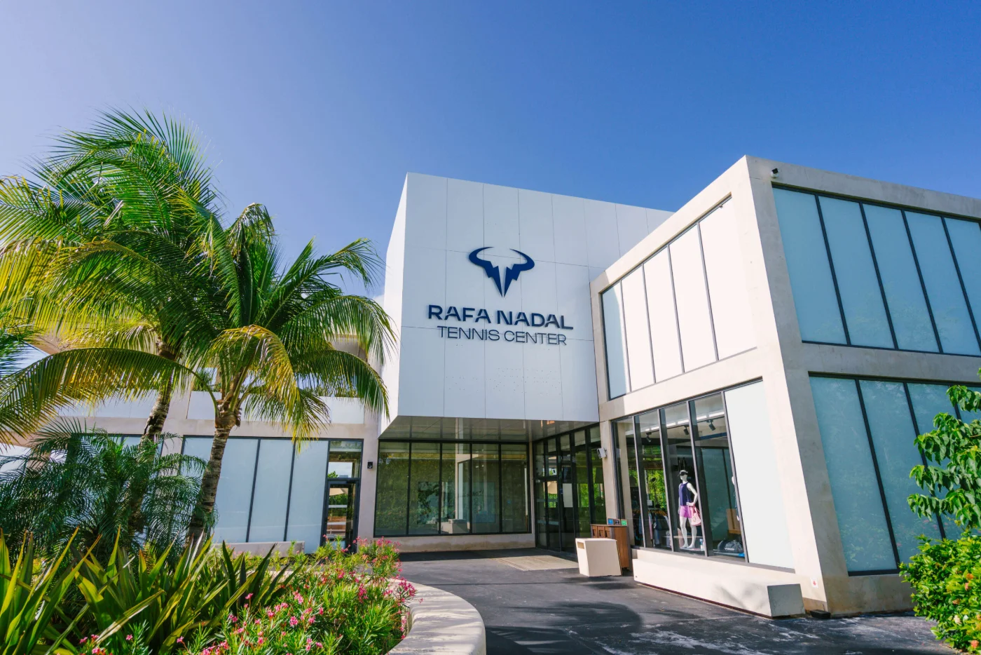 Entrance to the Rafa Nadal Tennis Center, the bull logo and the name on a white volume behind palms and flowering shrubs