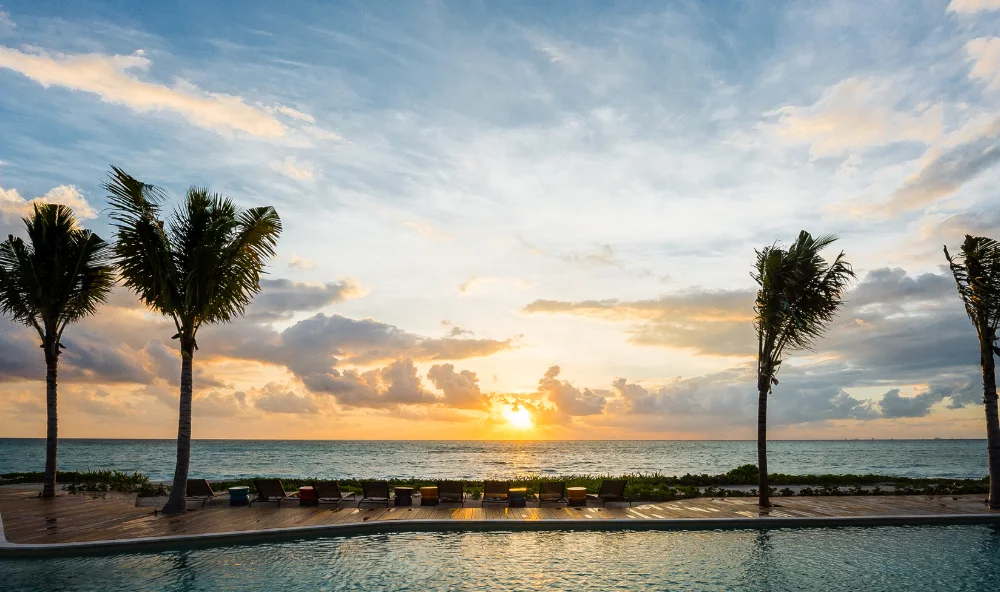 Stone and timber pergola at a Mayakoba hotel entrance at dusk, framing a view through palms to the Caribbean