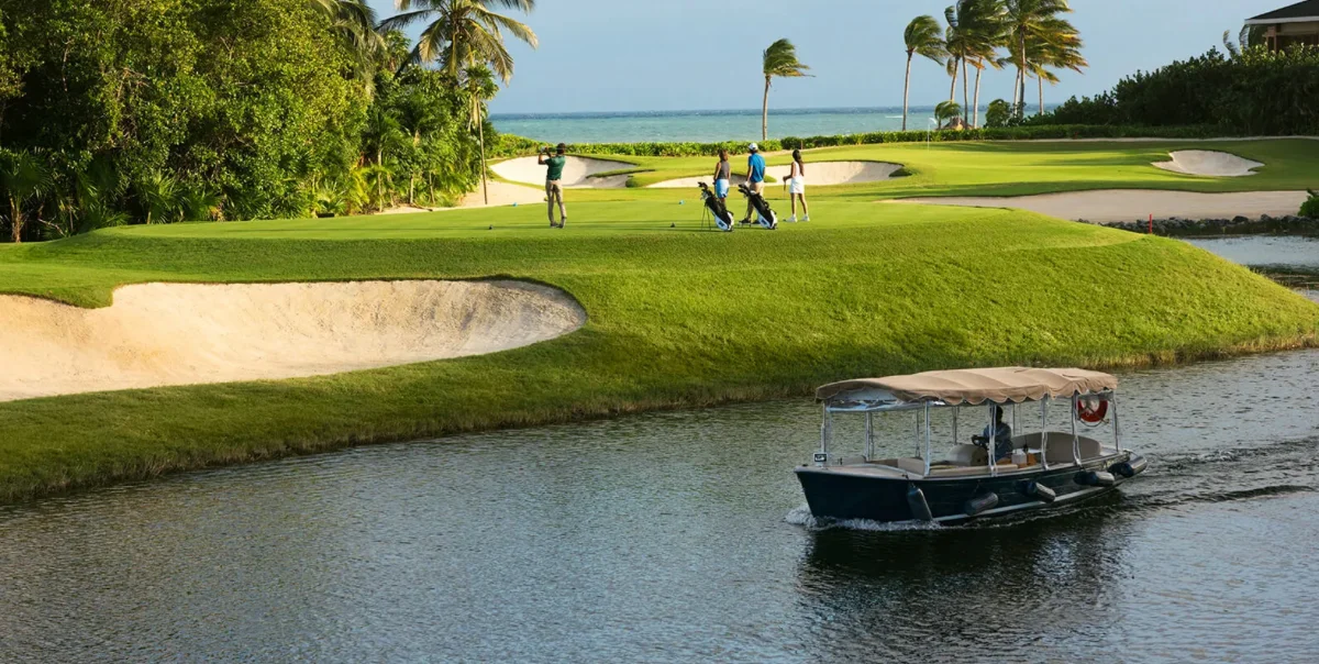 Golfers on a tee at El Camaleón while a covered boat passes along the canal below, the Caribbean behind