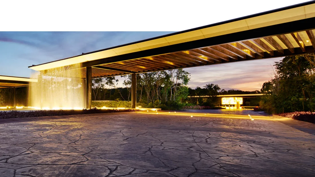 Muro de agua iluminado y cubierta de madera en el acceso de Ciudad Mayakoba al atardecer