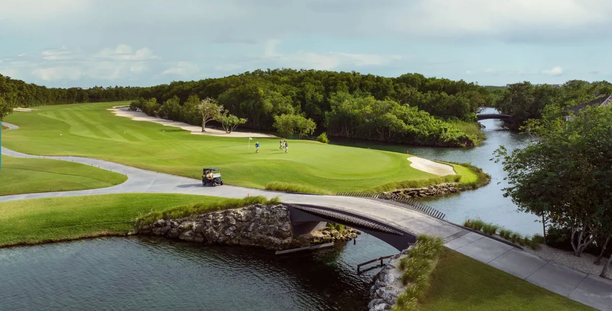 Pont de pierre franchissant un canal vers un green d'El Camaleón, golfeurs au putting et voiturette sur le chemin