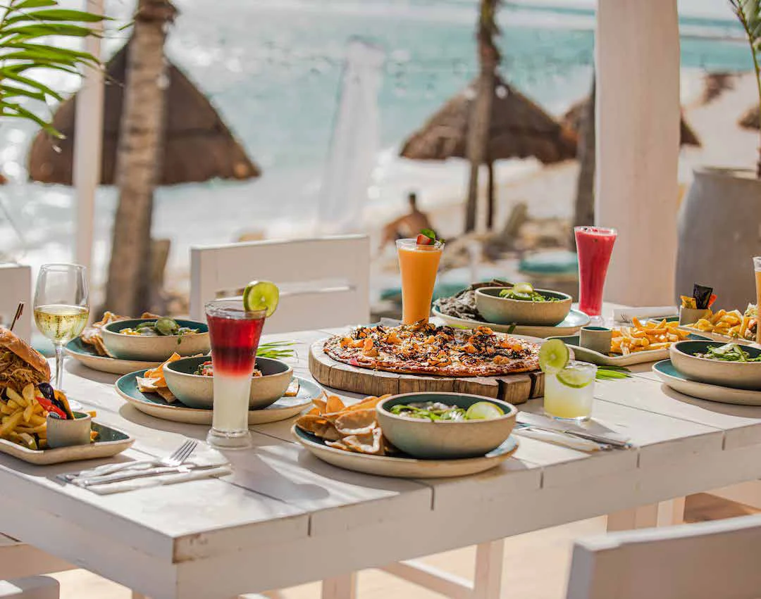 Table set with pizza, ceviche and drinks at Kay Beach Club, palapas and the sea behind